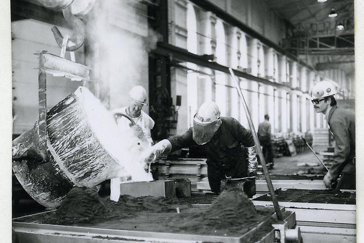 A black and white photo of a factory worker wearing a security helmet. He is pouring  hot liquid on a table. Another factory worker is watching.
