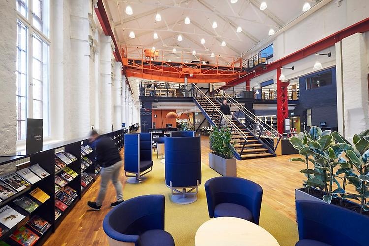 The lounge area with armchairs, tables and plants. Along the wall to the left, there is a long shelf with journals on display.