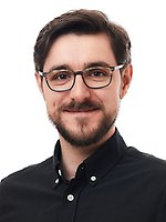 Man with glasses, short dark hair, and beard, wearing a black button-up shirt, smiling and facing forward against a white background.